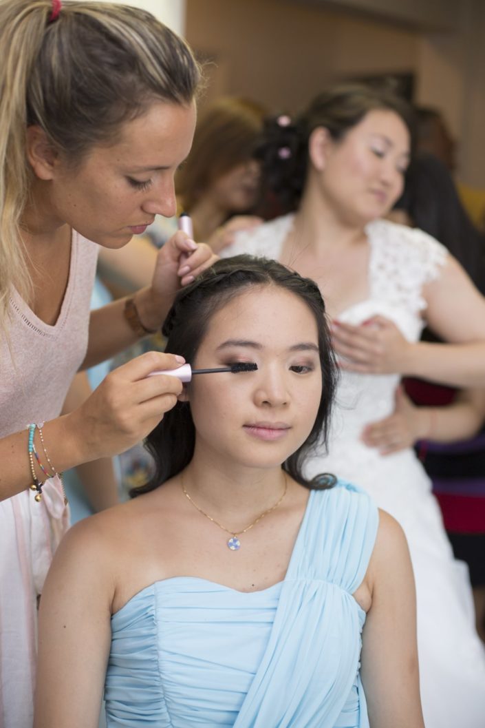 Maquillage d'une demoiselle d'honneur habillée en bleu, dans le fond la mariée, elle aussi asiatique, qui réajuste le bustier de sa robe blanche