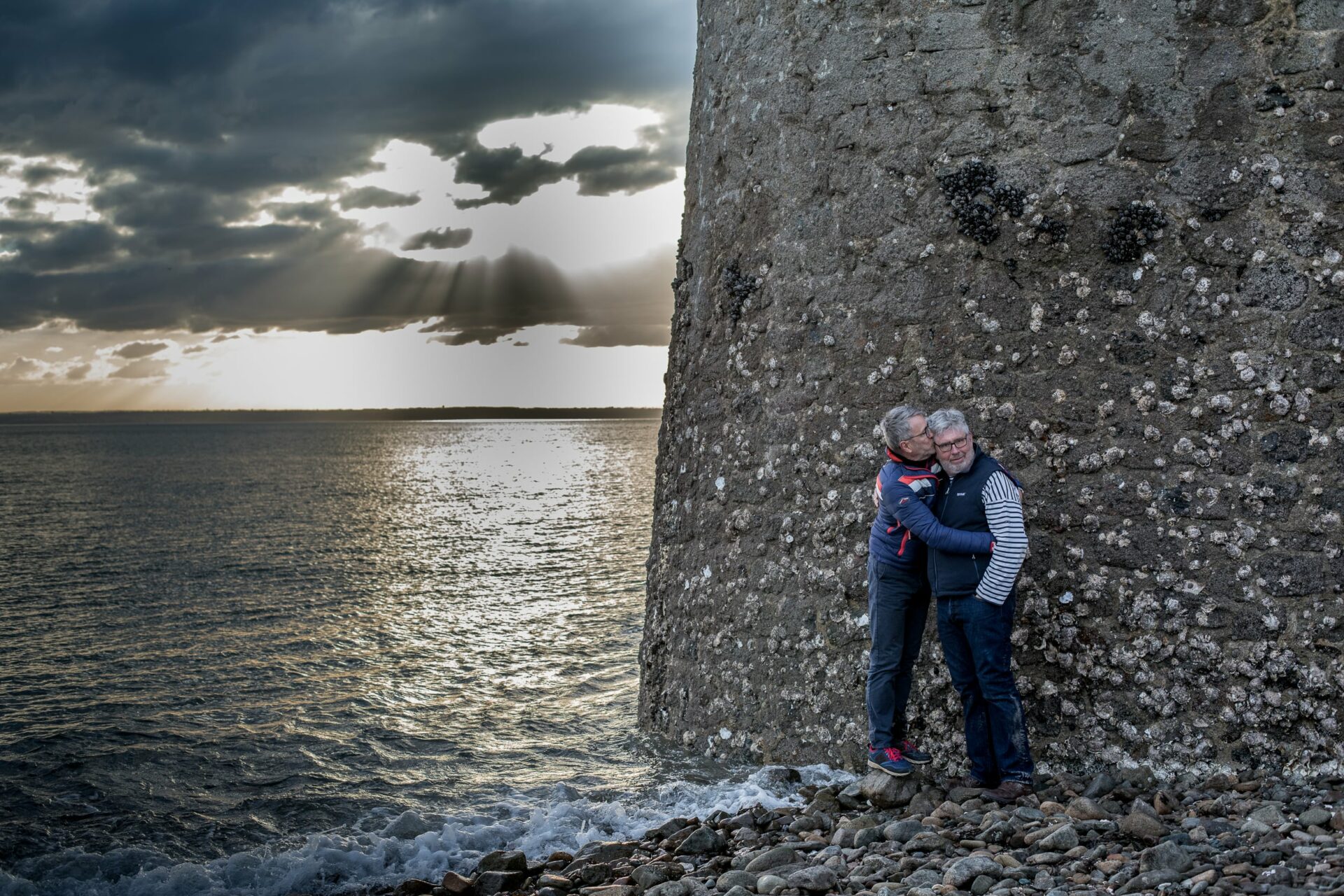 Fin de journée, mer bretonne au pied d'un phare dont on ne voit que le pied, 2 hommes enlacés tendrement dans cette immensité