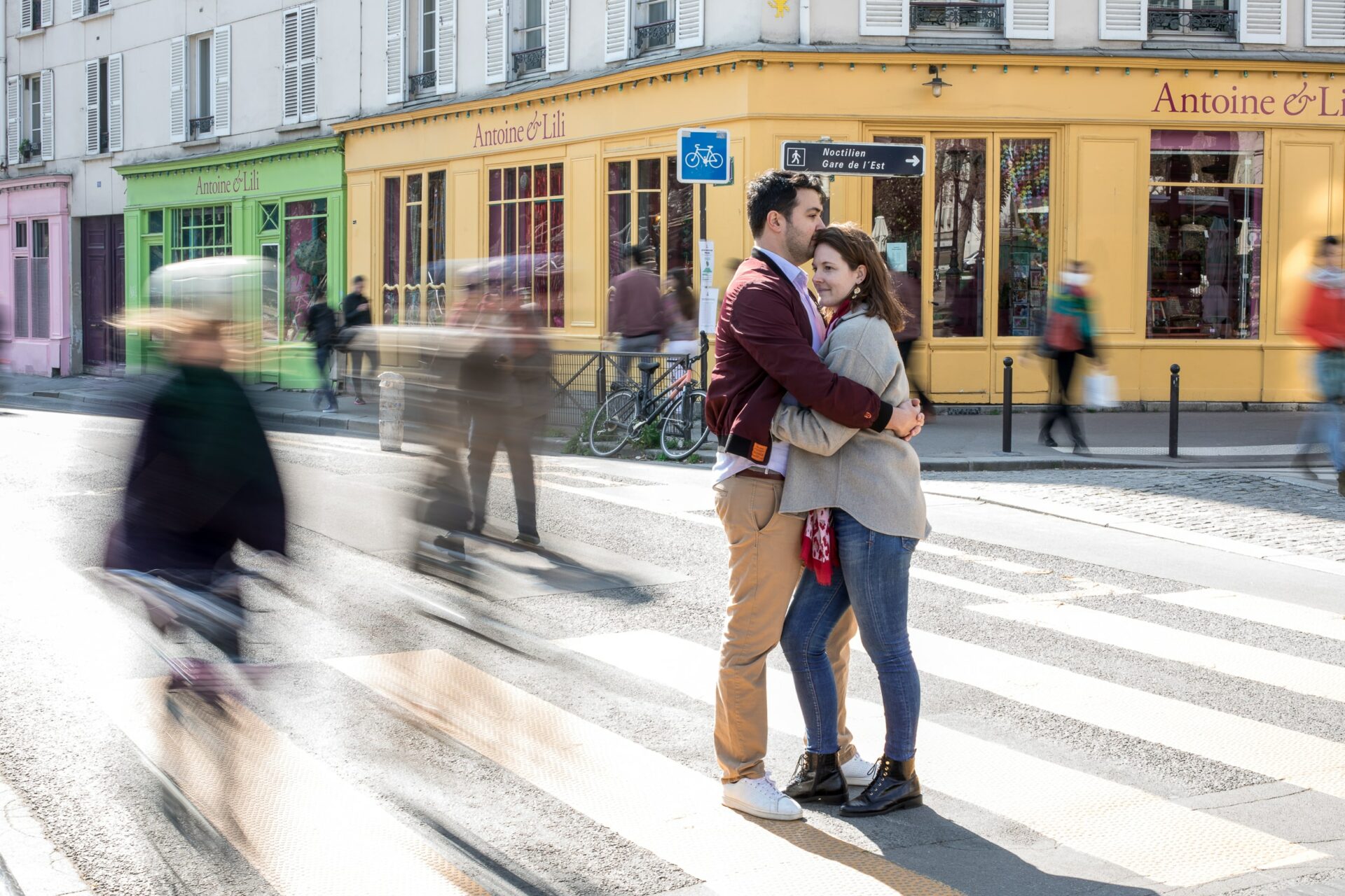 Au coeur de Paris, boutiques en couleur dans le fond, jaune, verte, rose, un couple serré l'un contre l'autre avec autour des fantômes de personnes qui filent